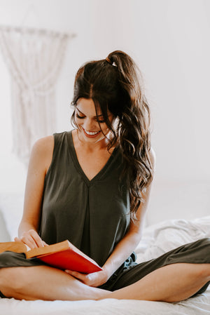 Model wearing olive colored Betty top from Jia Loungewear, sitting cross-legged on her bed reading a book and smiling.